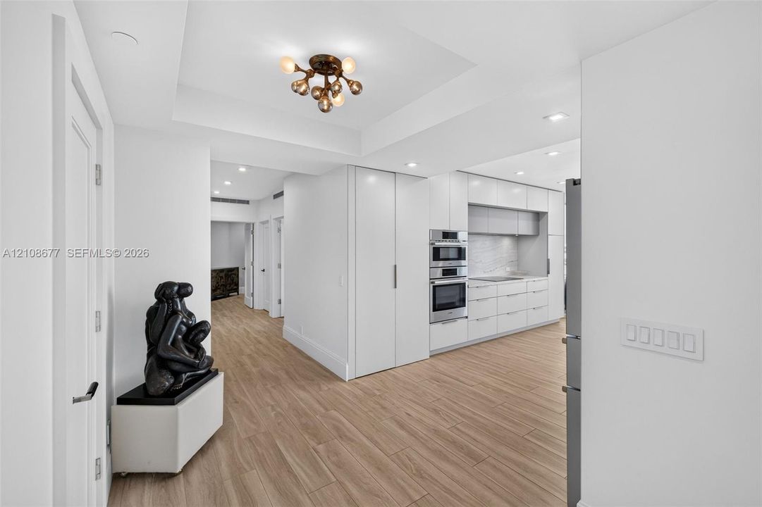 Widened hallway with primary bedroom straight down the hall, second bedroom next to it, plus gorgeous modern kitchen featuring white flat-panel cabinets, a gorgeous architectural multi-bulb flush-mount fixture, built-in Bosch stainless double ovens, and wood look plank porcelain flooring