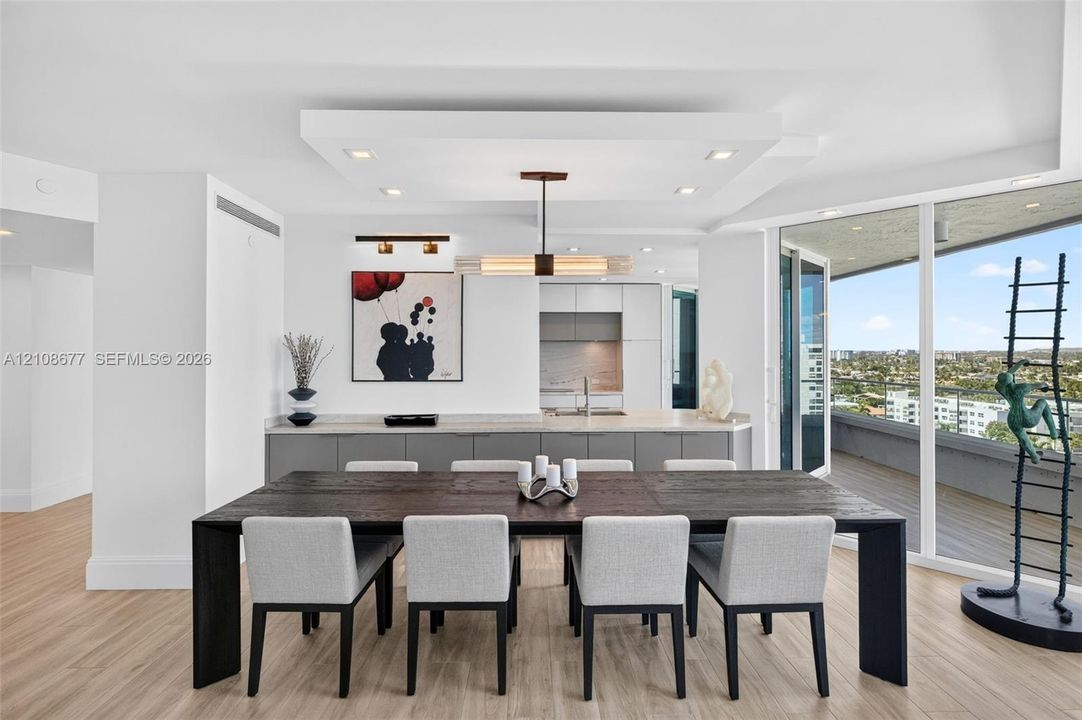 Open-concept kitchen featuring flat-panel cabinetry, light-toned stone counters, wood look plank porcelain flooring, tray ceiling with recessed lighting, and expansive floor-to-ceiling impact windows w/ sliders to the balcony. Primary bedroom down the hall on the left