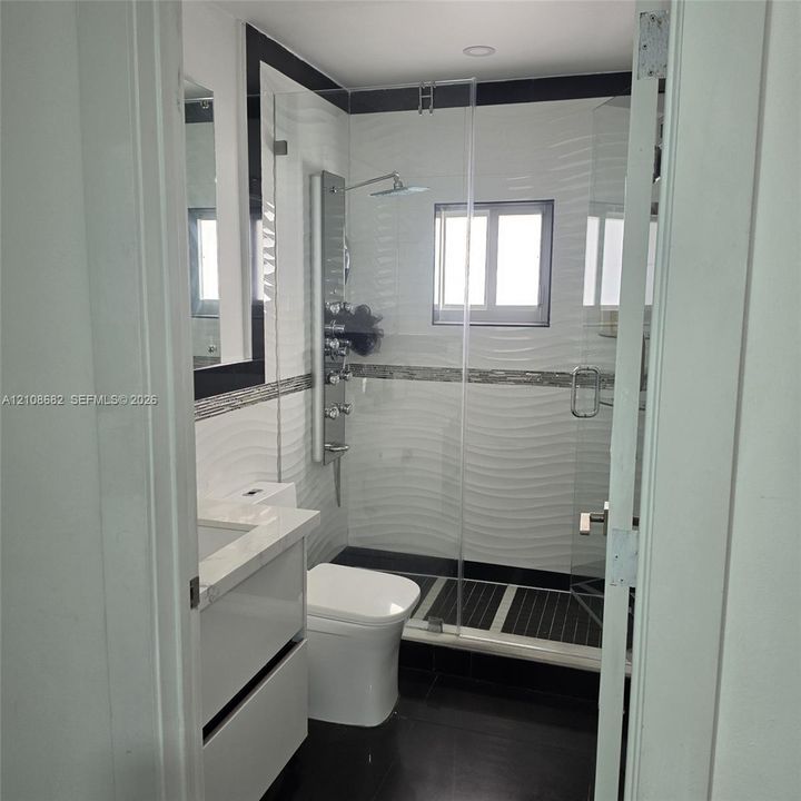 Bathroom featuring a wall-mounted shower panel system, glass shower enclosure, vanity with a stone-top counter, textured tile wall accents, and dark floor tiling