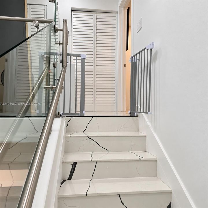 Staircase featuring white stone-finish treads with dark veining, stainless steel handrails, and a glass balustrade, complemented by white louvered closet doors