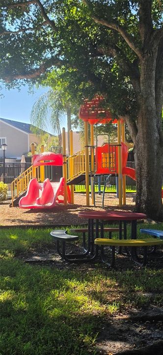 Outdoor common area featuring a multi-level play structure with slides, a surrounding black metal fence, mature canopy trees, and an anchored circular picnic table