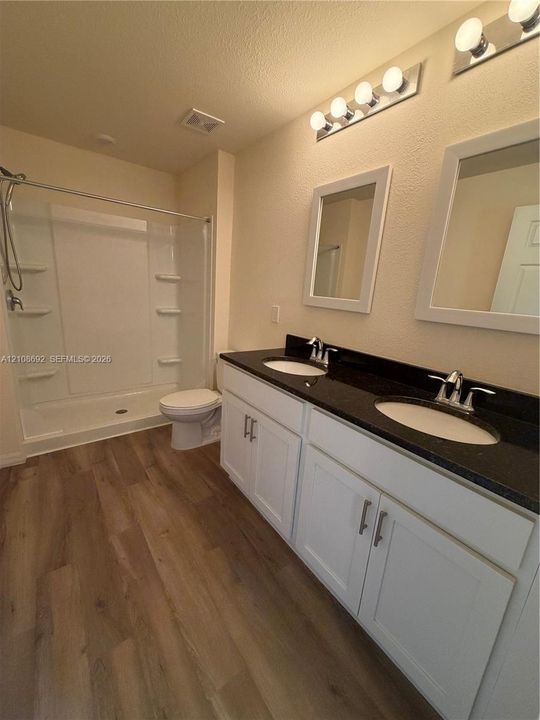 Dual-sink vanity with dark stone-finish countertop, white cabinetry, wood-finish flooring, and a shower-tub combination featuring built-in shelving