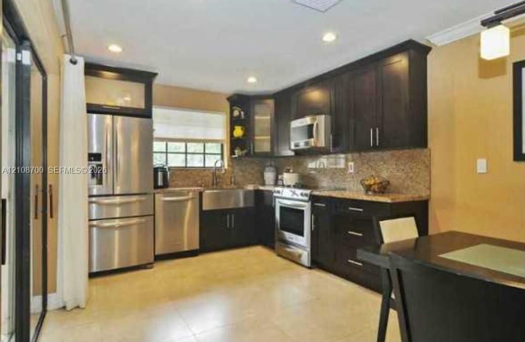 Kitchen featuring dark cabinetry, stainless steel appliances, stone-look countertops, farmhouse-style sink, and recessed lighting