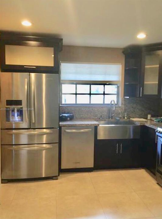 Kitchen featuring dark cabinetry with glass-panel accents, stainless steel appliances, farmhouse-style sink, stone-finish countertops, and recessed lighting