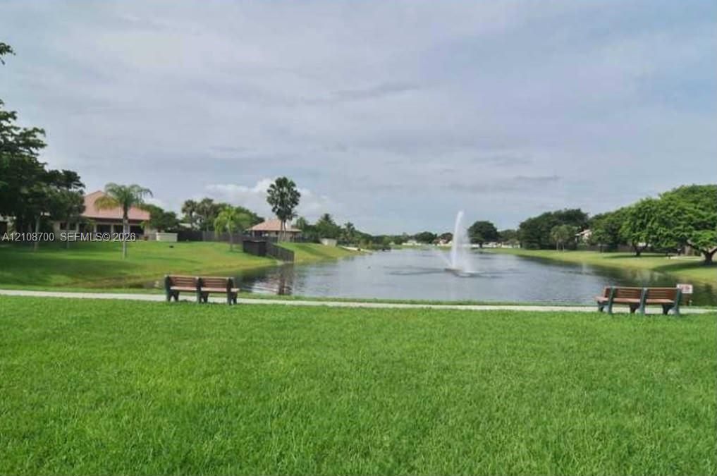 Waterfront common area featuring a central fountain, paved walking path, manicured lawn, and mature landscaping