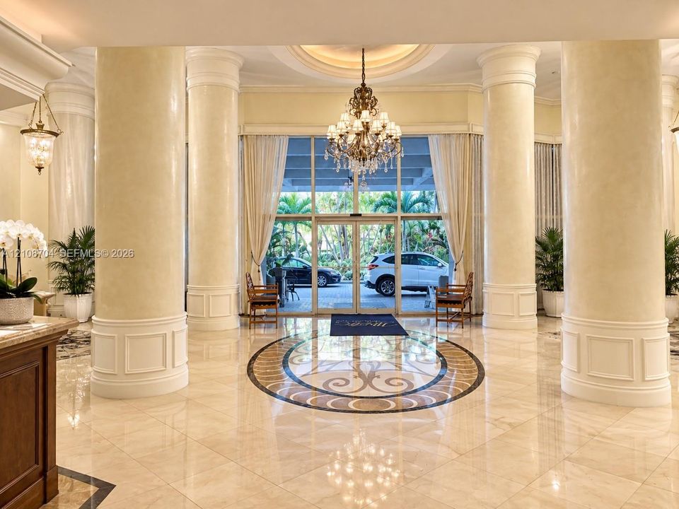 Grand lobby entrance featuring polished stone flooring with an inlaid mosaic medallion, structural fluted columns with detailed molding, a recessed circular ceiling medallion, and an ornate crystal chandelier
