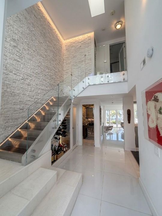 Staircase featuring a glass railing and integrated under-stair wine storage, accented by a floor-to-ceiling textured stone wall with cove lighting, large-format tile flooring, and a skylight