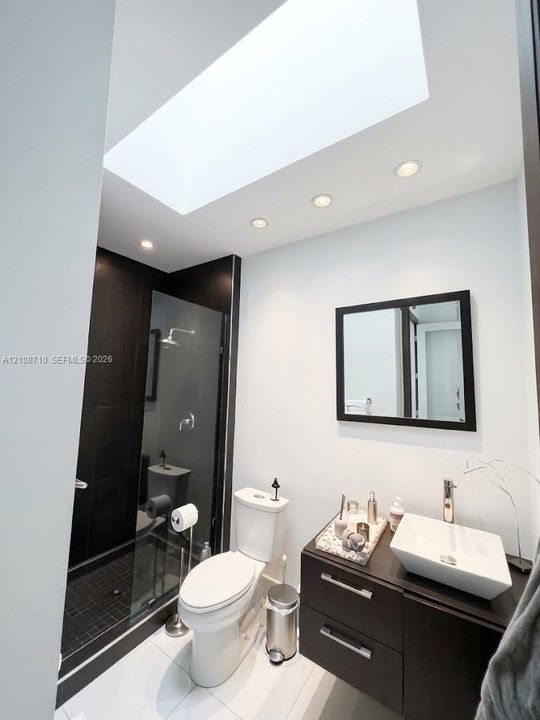 Skylight featuring recessed ceiling lighting, walk-in shower with dark tile surround, wall-mounted mirror, and vanity with vessel sink