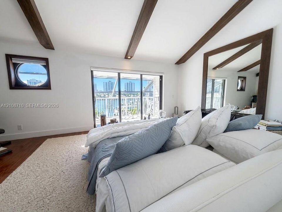 Vaulted ceiling with exposed wood-finish beams, sliding glass doors to a balcony, porthole-style accent window, large wall-mounted mirror with a wood frame, and wood-finish flooring