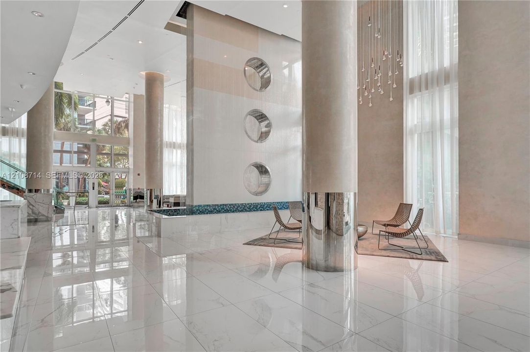 Grand lobby featuring expansive tile flooring, double-height ceilings, structural support columns with metallic accents, a decorative vertical wall feature with circular insets, and a suspended multi-light chandelier