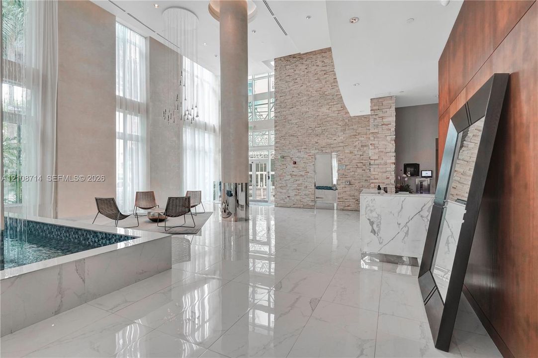 Grand atrium featuring floor-to-ceiling windows, polished stone flooring, a stacked-stone accent wall, a circular structural column, and a built-in water feature with mosaic tiling
