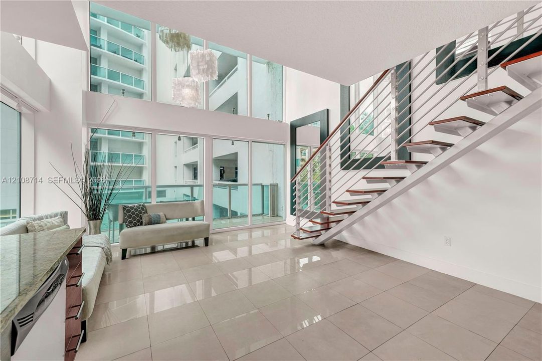 Double-height floor-to-ceiling windows, open-riser staircase with horizontal cable railing, neutral large-format tile flooring, and built-in stone-surface kitchen cabinetry