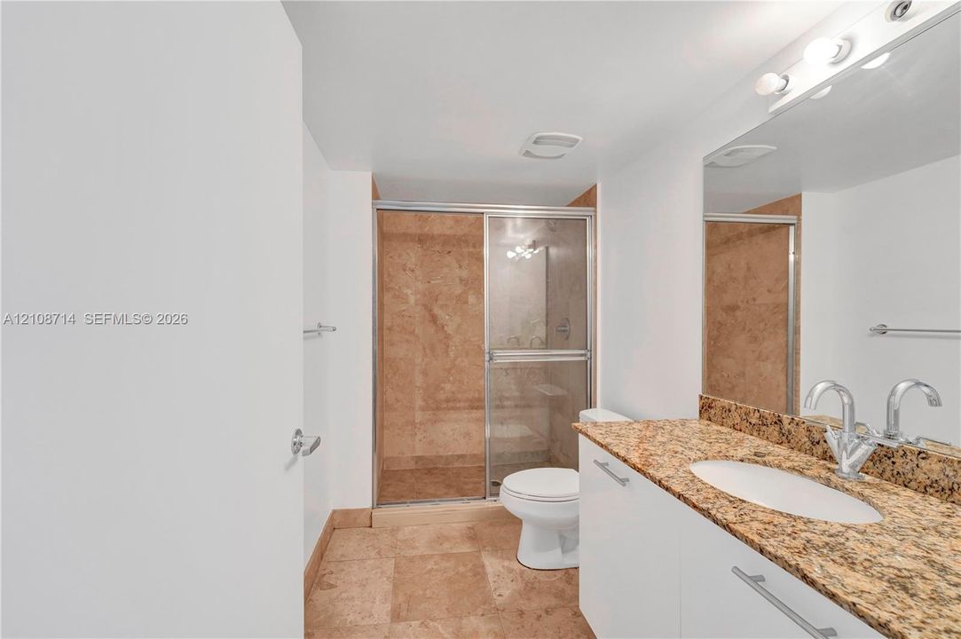 Bathroom featuring stone-finish floor tiling, a stone-topped vanity with an oval undermount sink, a large wall-mounted mirror, and a walk-in shower with stone tile surround and sliding glass doors