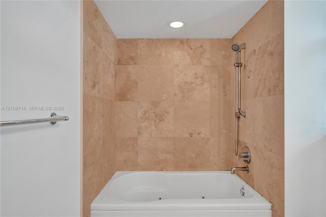 Built-in jetted tub featuring stone-tile surround, adjustable handheld shower fixture, metal faucet, and recessed lighting