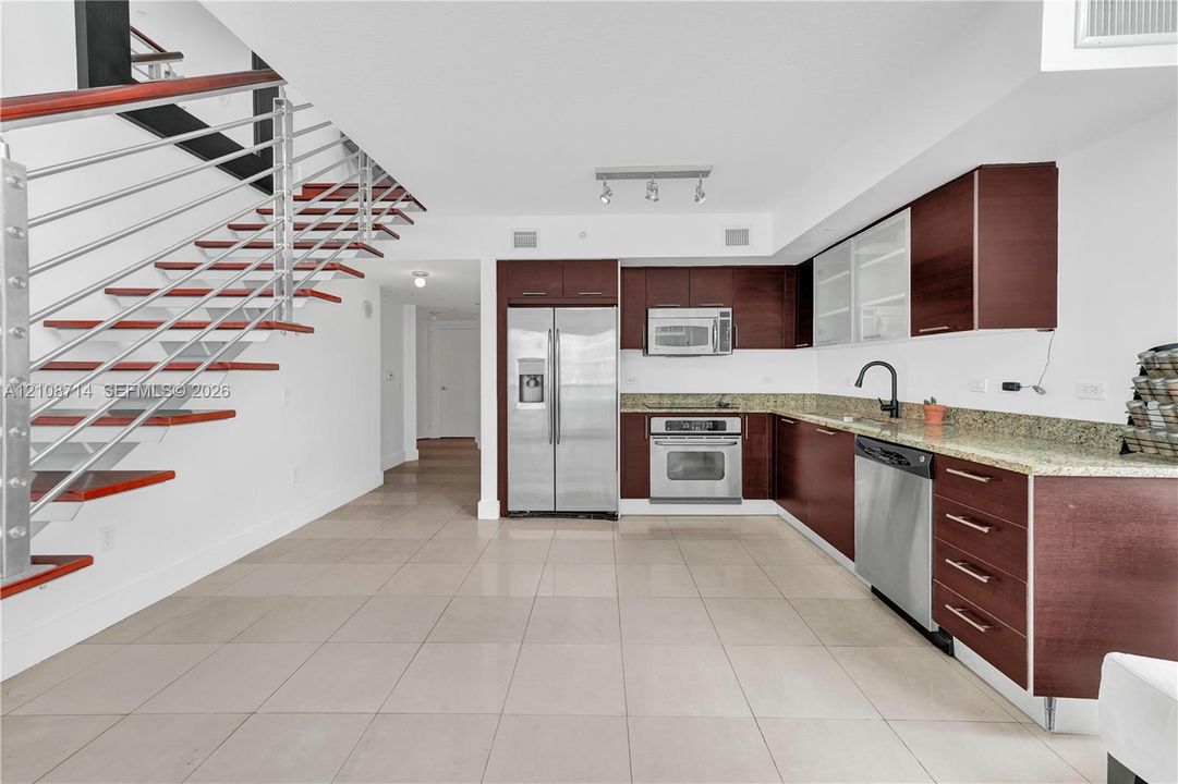 Kitchen featuring wood-finish cabinetry, stone countertops, stainless steel appliances, and large-format tile flooring, accented by an open-riser staircase with a horizontal cable railing system