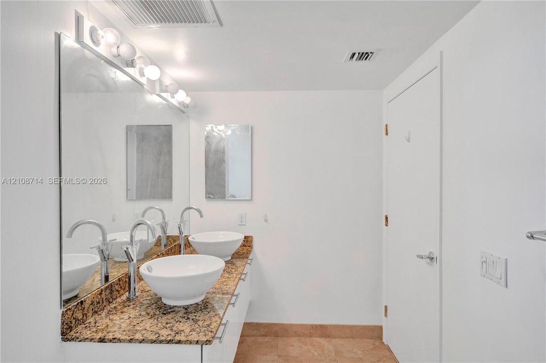 Double vanity featuring vessel sinks, stone-finish countertops, wall-mounted mirror, and multi-bulb lighting fixture