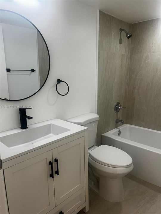 Bathroom featuring a white vanity cabinet with a rectangular basin, matte black faucet, circular wall-mounted mirror, and tile-surround bathtub