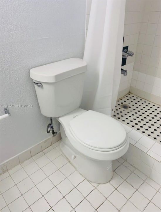 Bathroom featuring square white floor tile, a porcelain toilet, and a walk-in shower with black-and-white patterned mosaic flooring and a metal wall-mounted faucet assembly