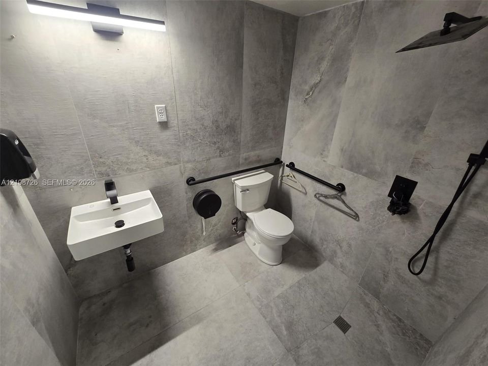 Large-format gray stone-finish tile wall and floor surfaces, wall-mounted white rectangular sink with matte black faucet, walk-in shower area with rainfall showerhead, and matte black hardware