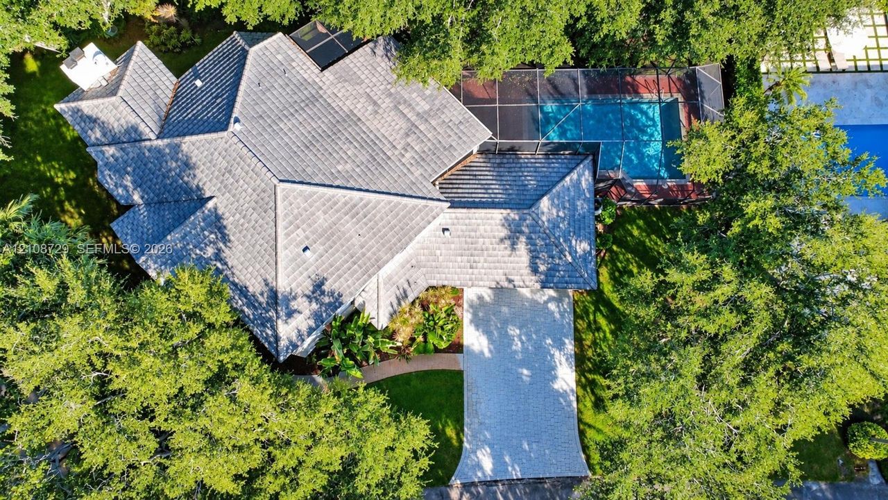 Aerial view featuring a multi-hipped roof, a screened-in swimming pool, paved driveway, and mature landscaping