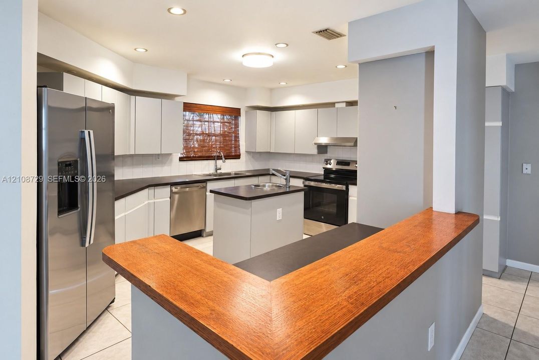 Kitchen featuring light-toned cabinetry, dark countertops, a center island with a built-in sink, stainless steel appliances, and a wood-finish breakfast bar