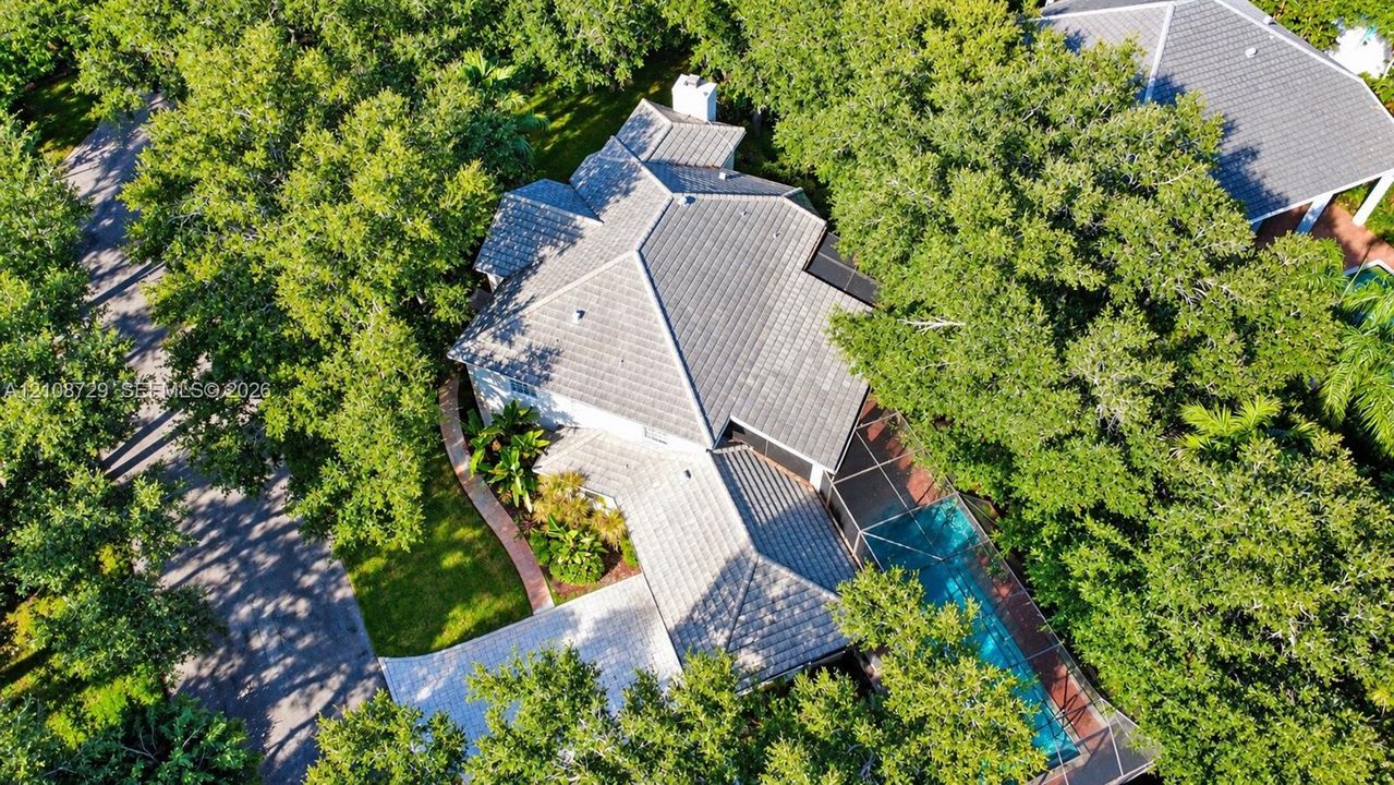 Aerial perspective of a multi-pitched tile roof residence featuring a screened-in swimming pool, paved driveway, brick walkway, and abundant mature trees