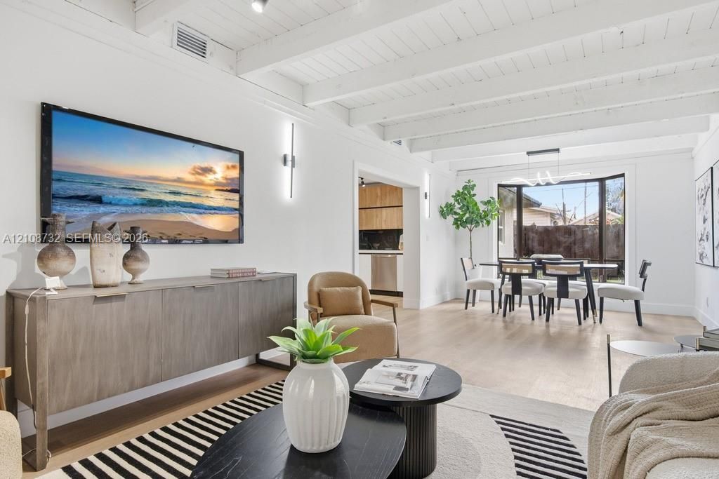 Exposed white-painted ceiling beams, wide-plank wood-finish flooring, linear wall-mounted lighting, and a bay window featuring a modern multi-light chandelier