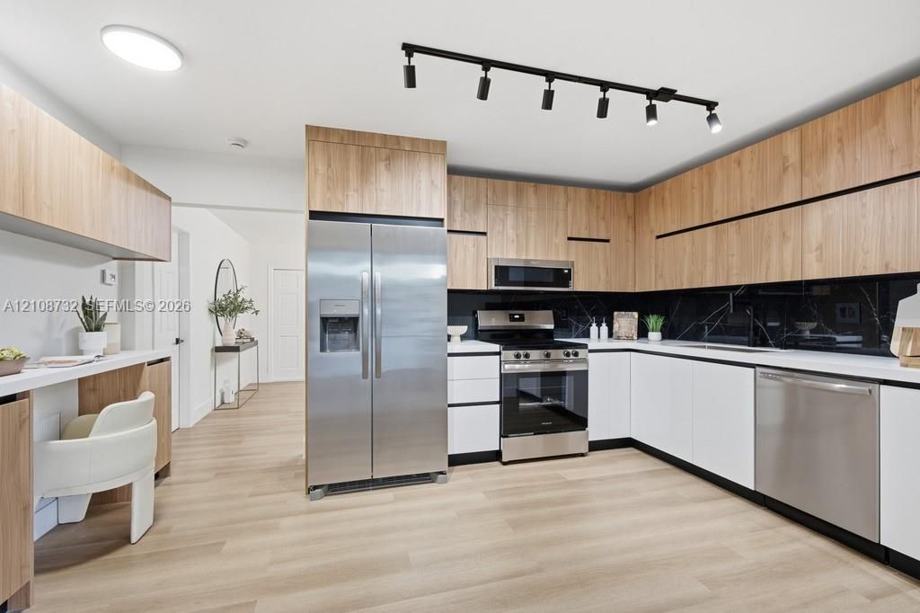 Kitchen featuring flat-panel wood-finish cabinetry, stainless steel appliances, dark stone-finish backsplash, built-in desk area, and matte black track lighting