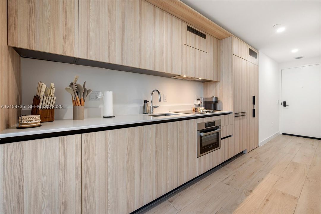 Modern kitchen featuring light wood-finish cabinetry, solid surface countertops, built-in stainless steel appliances, and wide-plank wood-finish flooring