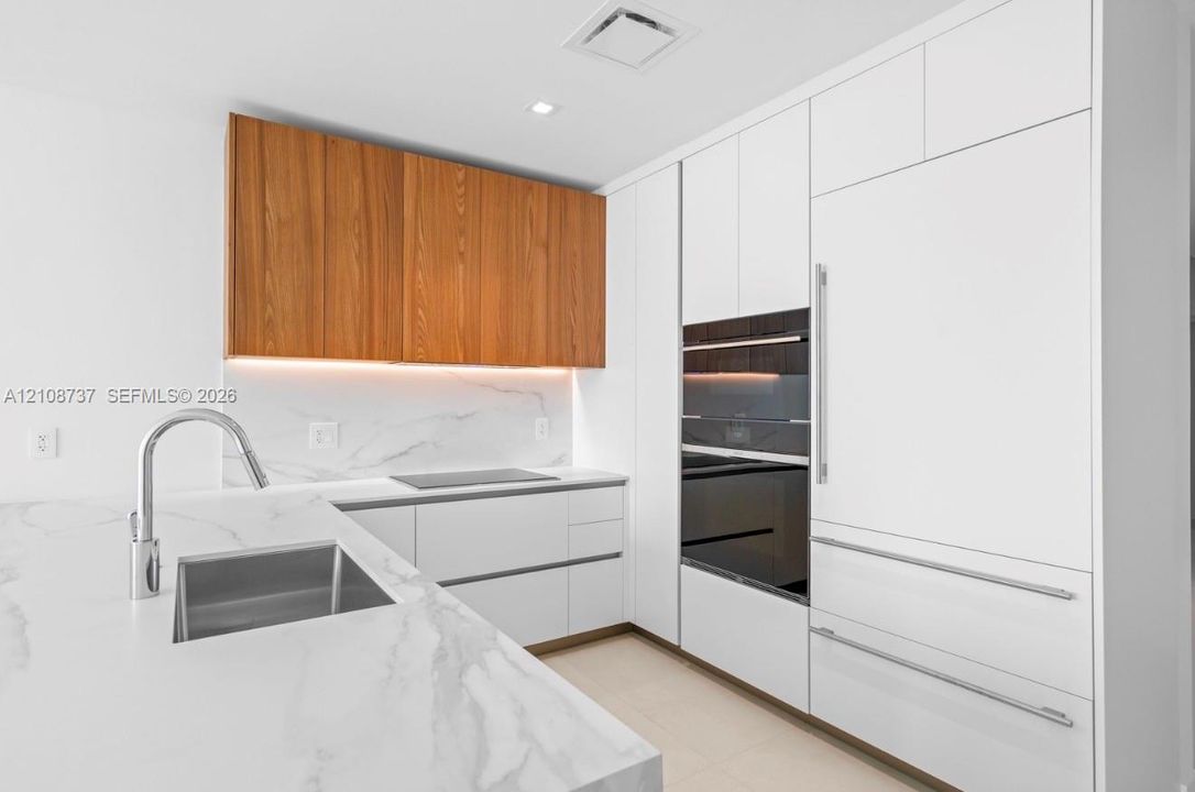 Modern kitchen featuring white flat-panel cabinetry, wood-veneer upper cabinets, light-toned stone-finish countertops and backsplash, integrated stainless steel appliances, and an undermount rectangular sink with a gooseneck faucet