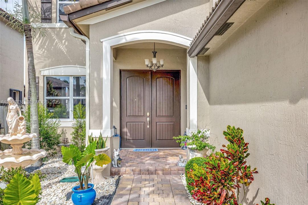 Covered entryway featuring arched architectural detailing, double-door entrance, stone-paved walkway, and a multi-tiered tiered fountain
