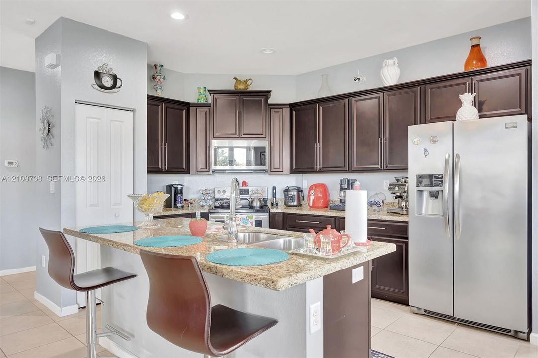 Kitchen featuring dark wood cabinetry, stone countertops, stainless steel appliances, a central island with a double-basin sink, and recessed ceiling lighting