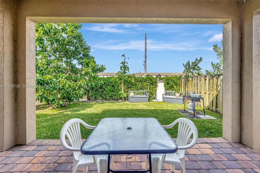 Covered patio featuring paver flooring, overlooking a backyard with a manicured lawn, mature trees, and a wooden privacy fence