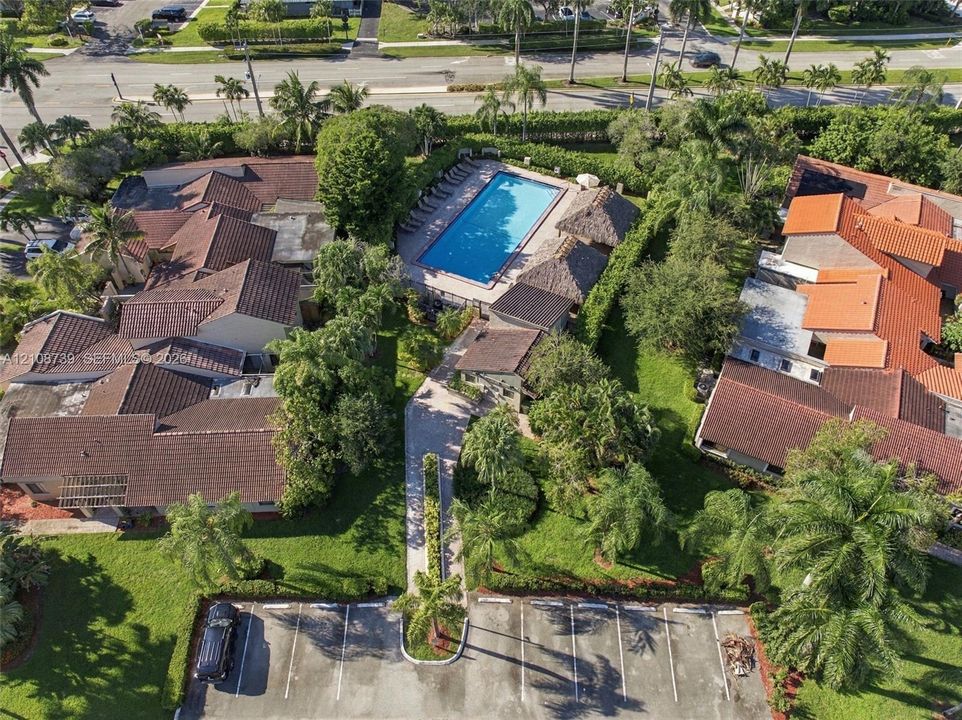 Aerial perspective of the property featuring a rectangular swimming pool, thatched-roof structures, concrete paved walkways, asphalt parking area, and mature landscaping