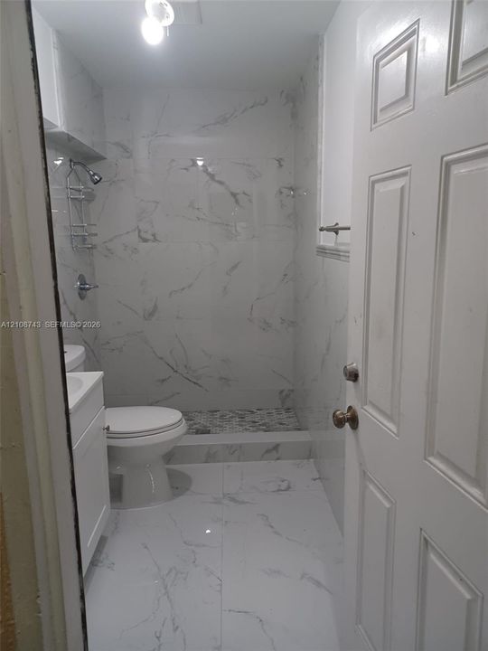 Full-height tile surrounds with stone-pattern finish, walk-in shower with mosaic tile floor, white vanity cabinet, ceramic toilet, and six-panel interior door