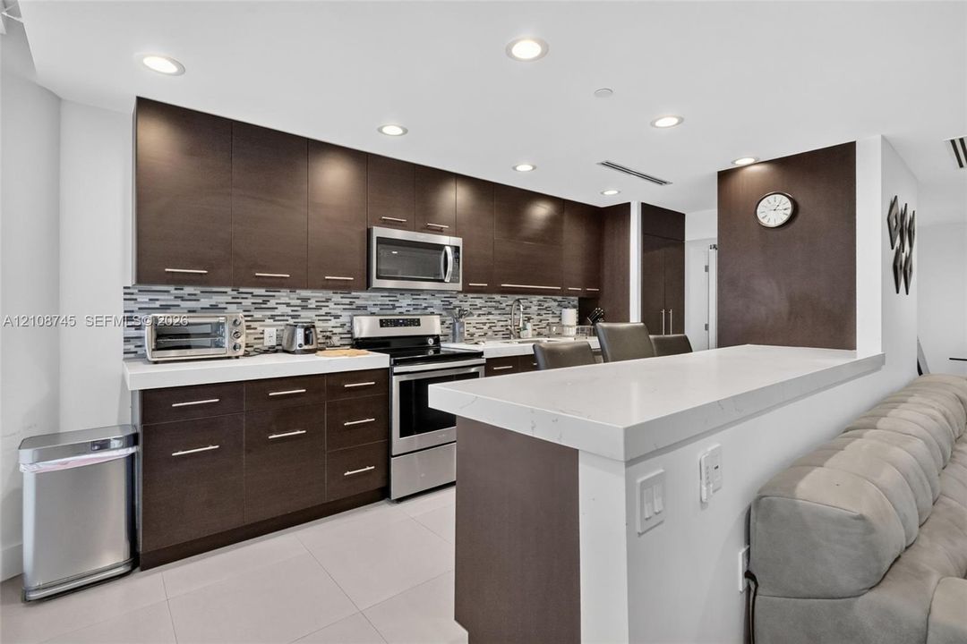 Kitchen featuring dark wood-finish cabinetry, solid surface countertops, a mosaic tile backsplash, stainless steel appliances, and recessed lighting