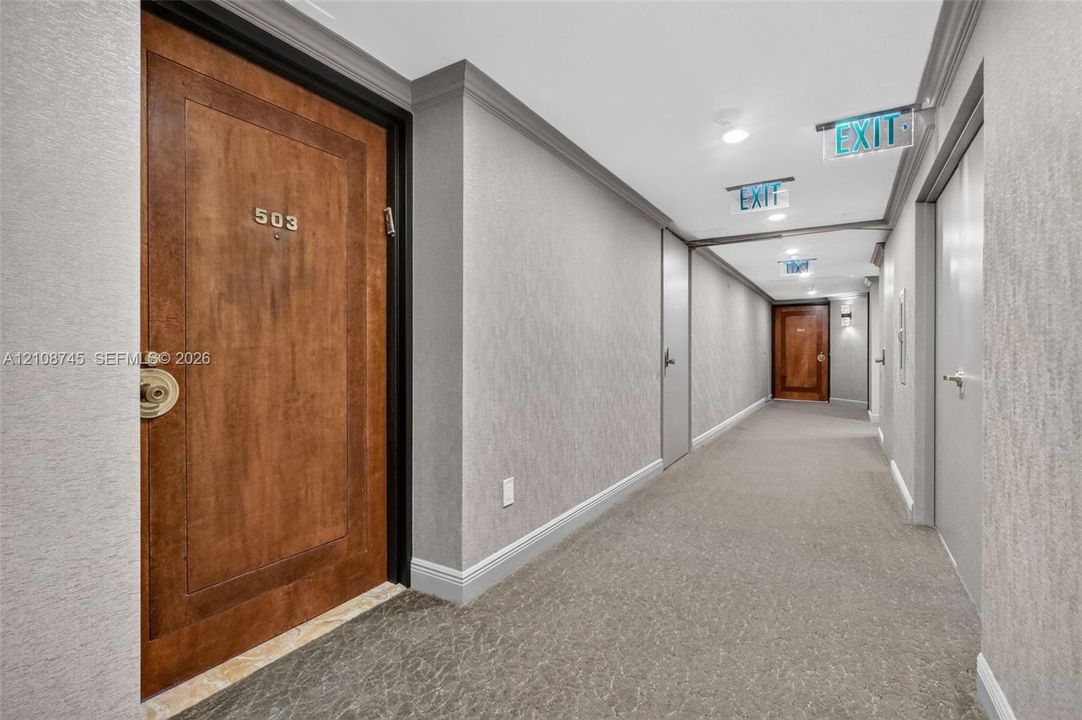 Interior hallway featuring neutral textured wall coverings, crown molding, patterned carpeting, and solid wood-finish entry doors with metallic hardware