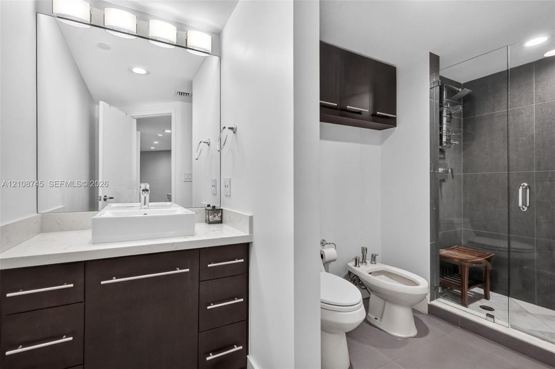Bathroom featuring a vanity with quartz countertop and vessel sink, wall-mounted cabinetry, bidet, and a glass-enclosed shower with dark tile surround