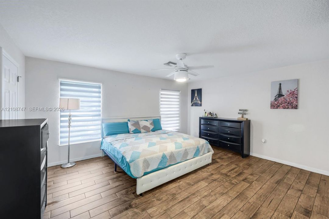 Wood-finish flooring, neutral wall color, tiered light-filtering window shades, and a ceiling-mounted light fixture with a fan