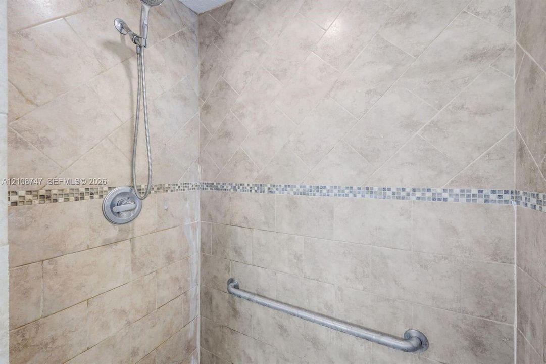 Shower stall featuring diagonal tile wall patterns, horizontal stone tile accents, a handheld shower fixture, and a wall-mounted support bar