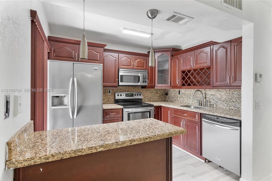 Kitchen featuring cherry-finish cabinetry with built-in wine storage, stone-patterned countertops, stainless steel appliances, and pendant lighting