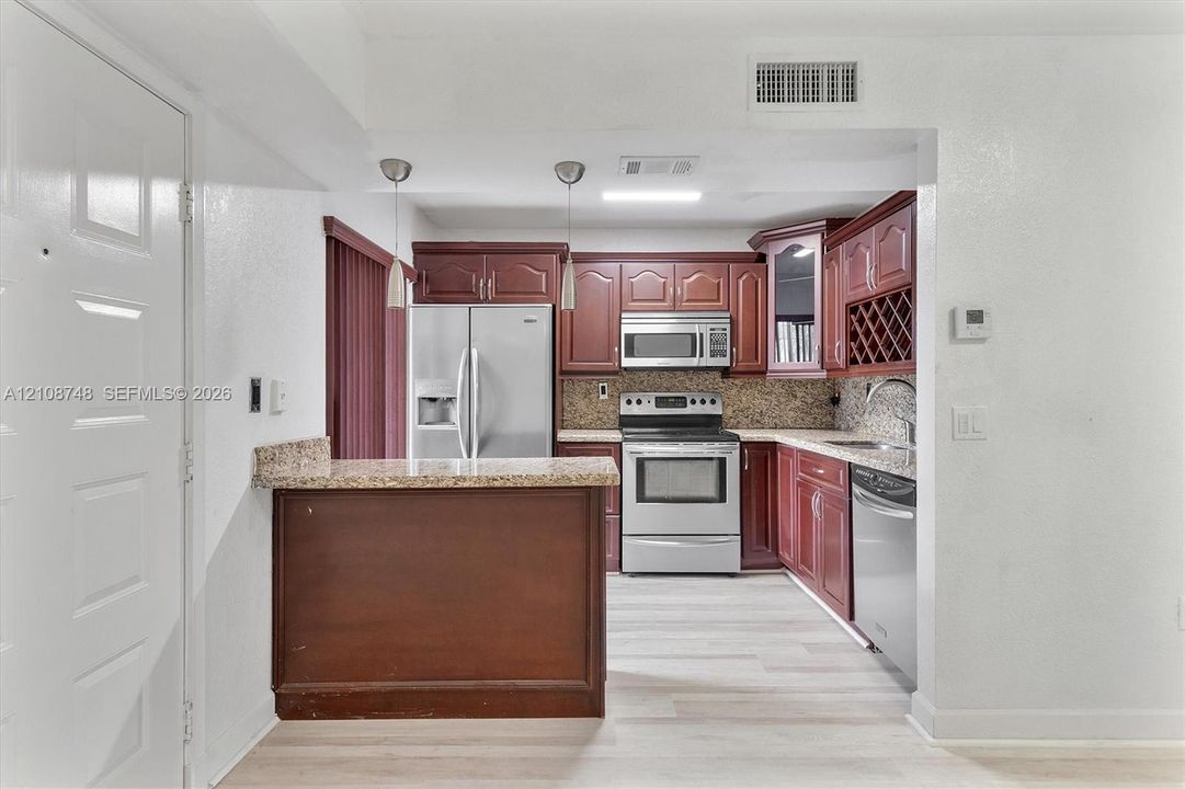 Kitchen featuring wood cabinetry, stone-finish countertops, stainless steel appliances, a peninsula with pendant lighting, and light-tone wood-finish flooring