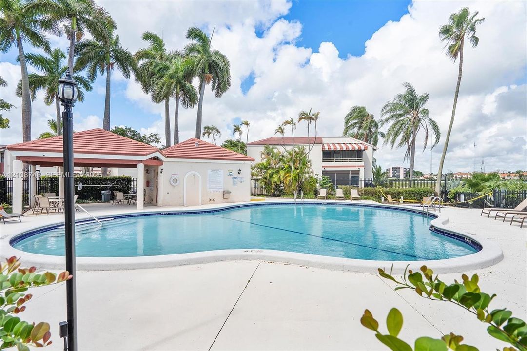 Curved swimming pool with a concrete pool deck, covered pavilion featuring a terracotta-style tile roof, and mature palm trees