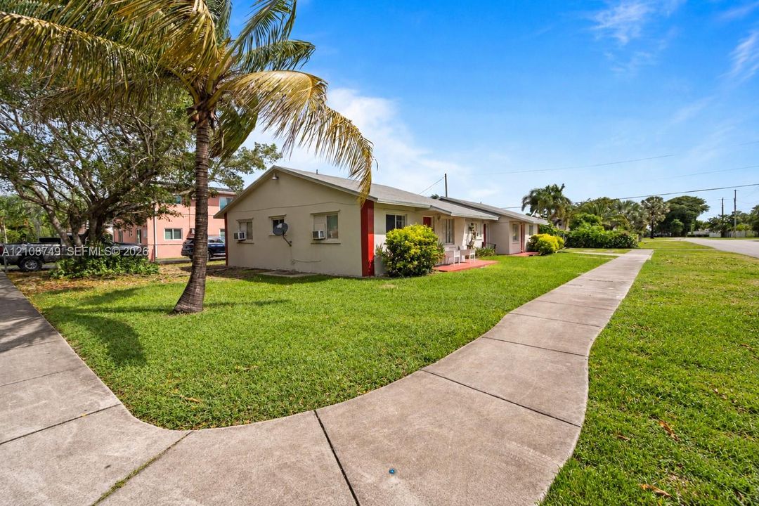 Exterior view featuring a single-story structure with a light-colored stucco finish, contrasting red trim accents, a concrete walkway, and established landscaping including mature palm trees