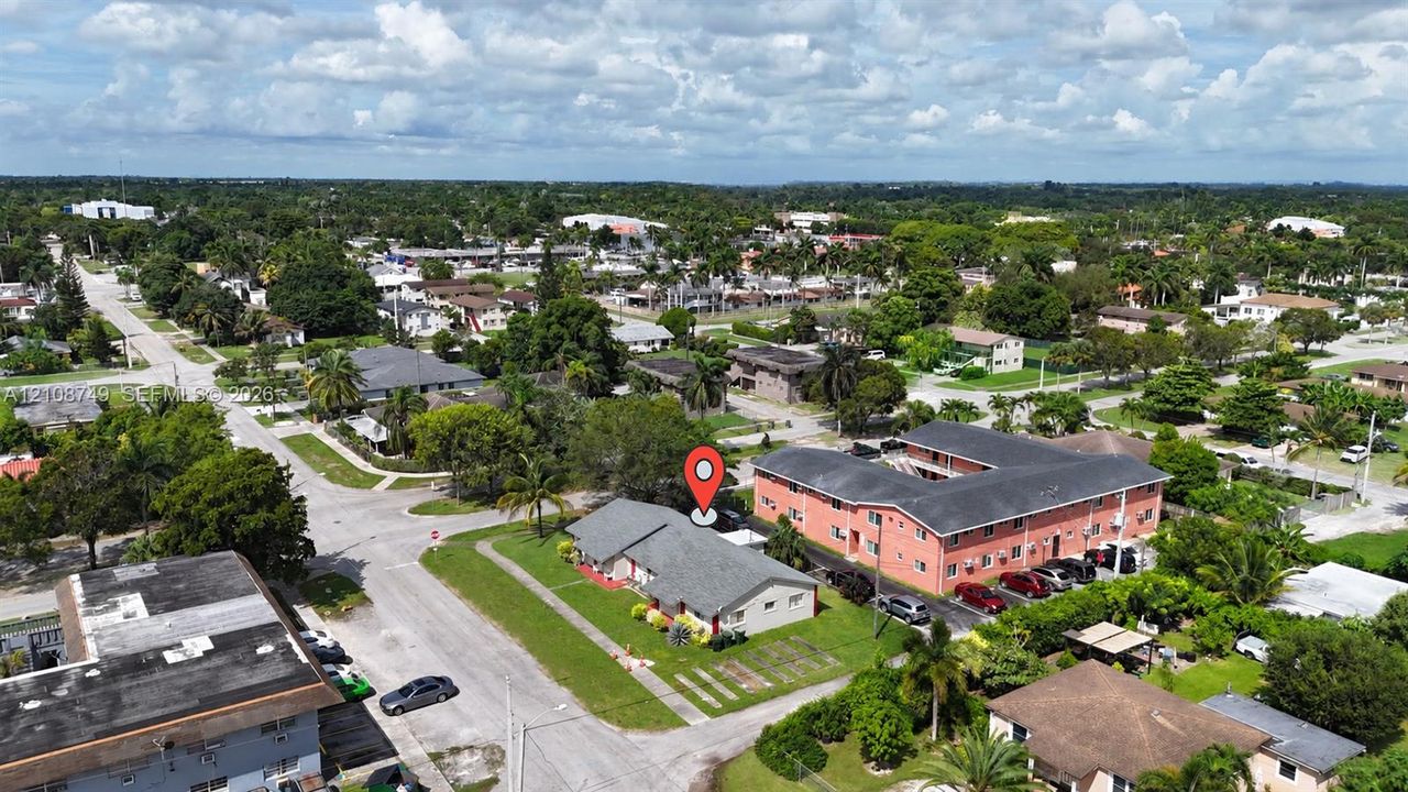 Aerial perspective of a single-story structure with a pitched shingle roof, positioned on a corner lot with established green space and mature palm trees