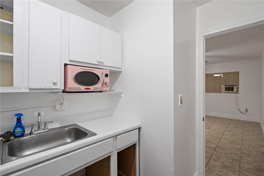 Kitchenette area featuring white cabinetry, a single-basin stainless steel sink, and light-toned solid surface countertops, featuring tile flooring in the room beyond