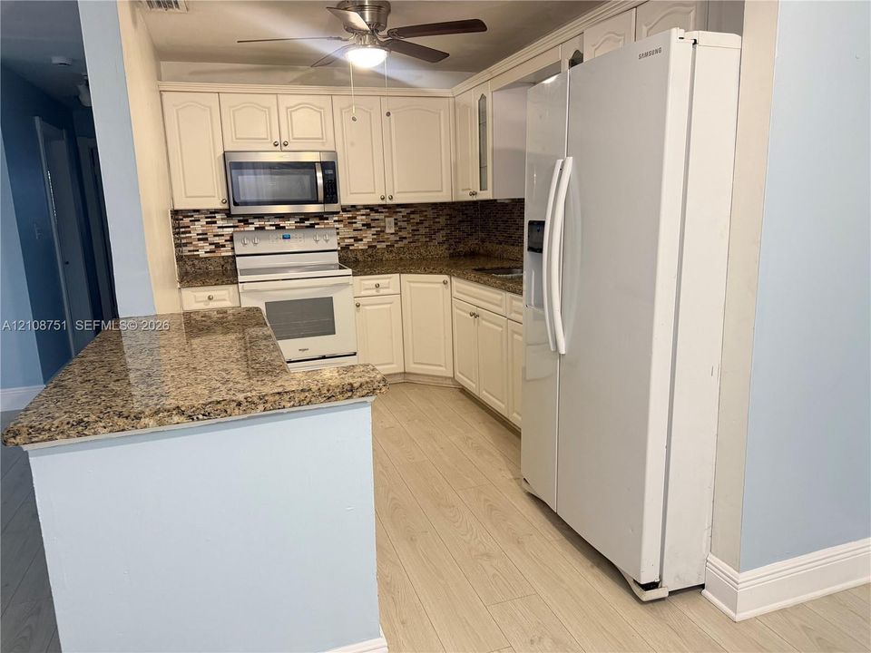 Kitchen featuring cream-toned cabinetry, stone-surface countertops, a multicolored mosaic tile backsplash, wood-finish flooring, and a ceiling-mounted light fixture with integrated fan