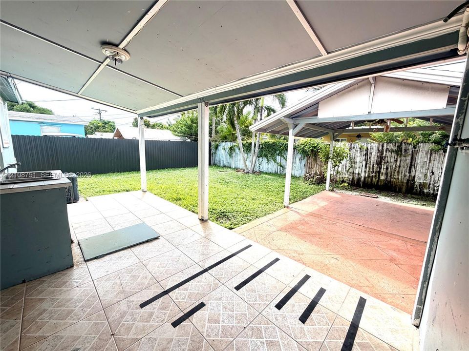 Covered outdoor patio featuring tile flooring, built-in counter with sink, and metal roof structure overlooking a fenced backyard with concrete patio area