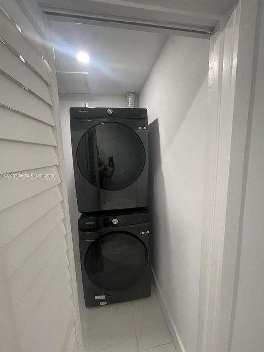 Stacked, front-loading laundry unit in a dedicated closet featuring louvered doors and neutral floor tiling