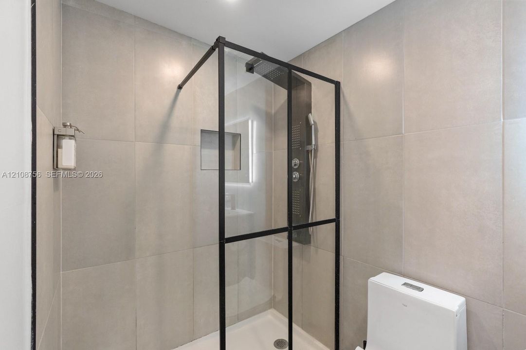 Walk-in shower enclosure featuring matte black framing, floor-to-ceiling large-format tile surround, integrated shower panel with handheld sprayer, recessed wall niche, and low-profile white base